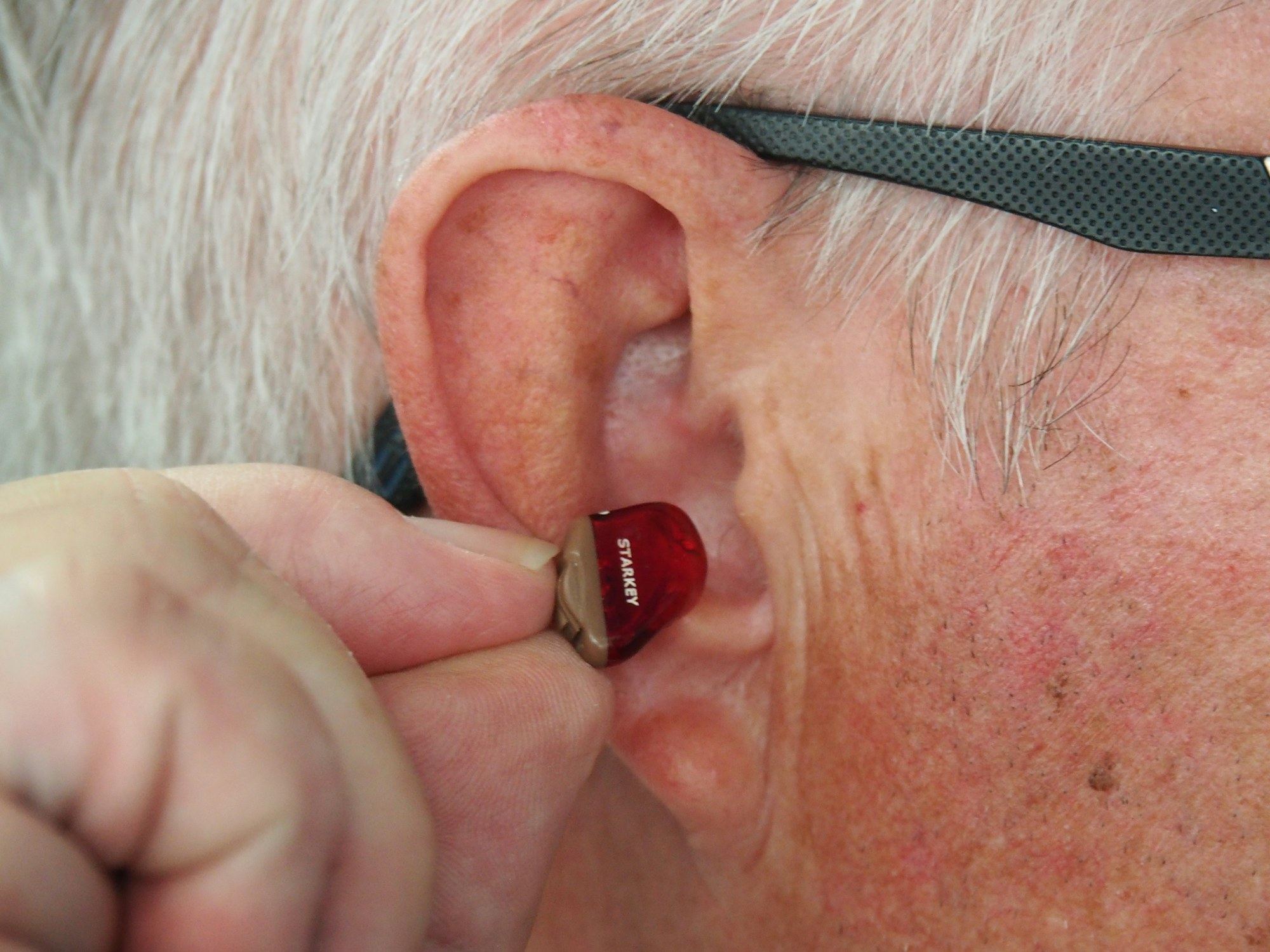 A hand inserts a small hearing aid into an elderly person's ear.